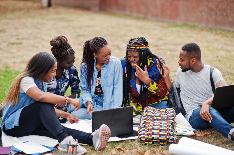 Group of five african college students spending time together on campus at university yard. Black afro friends sitting on grass and studying with laptops.